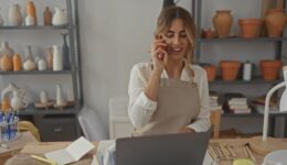 Young woman holding phone to ear in pottery studio while checking orders and reaching for clay tools; creativity focus serenity.