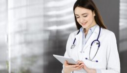 Cheerful intelligent woman-doctor is holding a tablet computer in her hands, while she is standing in a clinic. Portrait of a friendly physician woman at work. Perfect medical service in a hospital. Medicine concept