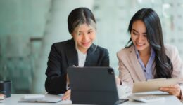 Two Asian business women working together using digital tablets discussing new startup project ideas. Analyze the planning and statistics of the financial and investment markets at the office.