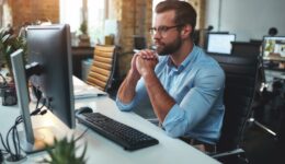 Thinking. Side view of young bearded man in eyeglasses and formal wear working on computer and keeping palms pressed together while sitting in the modern office