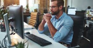 Thinking. Side view of young bearded man in eyeglasses and formal wear working on computer and keeping palms pressed together while sitting in the modern office Thinking. Side view of young bearded man in eyeglasses and formal wear working on computer and keeping palms pressed together while sitting in the modern office