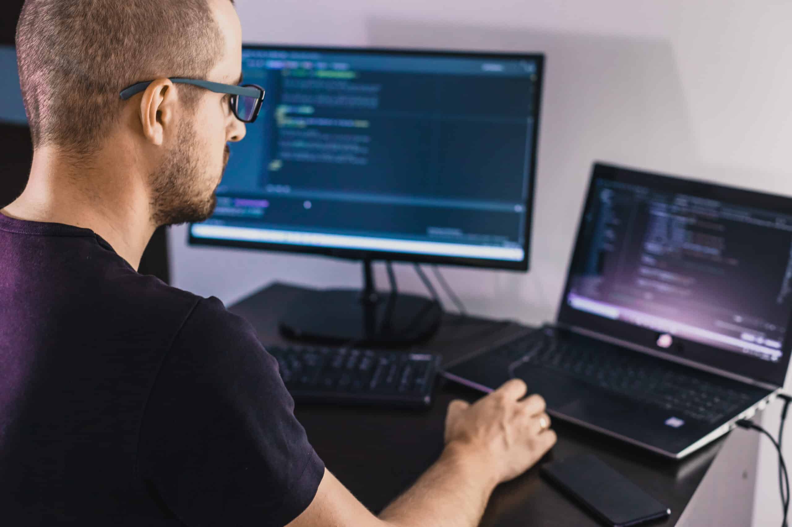 Computer programmer in glasses. Information Technology. Women in business. The guy works from home. IT professional managing SQL scripts on dual monitors during an automated database migration, showcasing advanced database migration services.